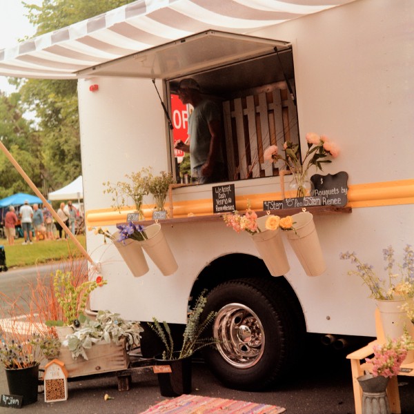 Flower Truck A white trailer with an open sale window with a striped awning above it, a person can be visible inside looking out the open back. The window has a ledge with cans hanging off of it holding flower stems and signs advertising the prices per stem ranging from $2 to $8. There are also pots with flowers and plants on the ground outside the trailer.