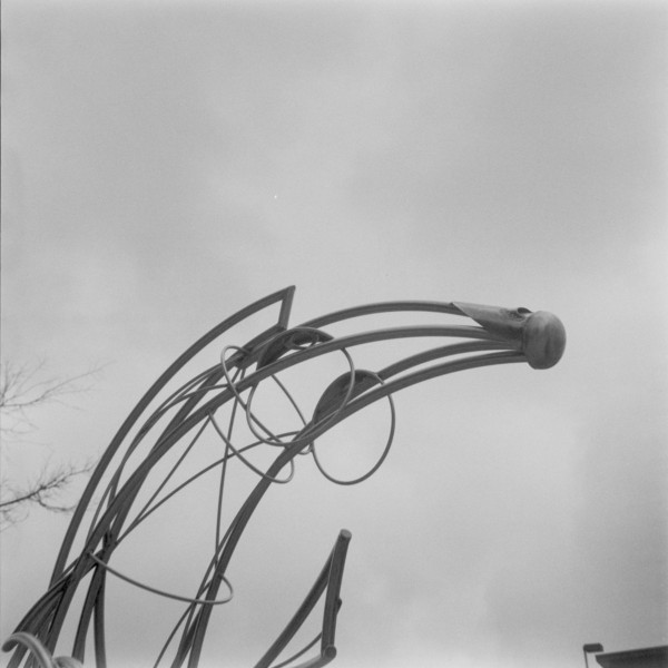 Sculpt Bulb Black and white photo of a metal sculpture of an abstract bee stinger (open to interpretation) made of pipes and circles that are capped off in a bulb, against a stark and gray sky.