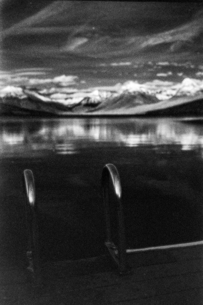 A black and white photo with a pair of raised ladder handles attached to the end of a dock which is sitting on a lake that stretches to the out of focus background framed by mountains with snowy peaks under a darker sky with swirly white clouds. Shot with a Voightlander BessaR2 and 7Artisan35f2 lens, on RolleiInfrared, and developed with Rodinal by Shom Bandopadhaya. Licensed under Creative Commons Attribution-NonCommercial-ShareAlike (CC BY-NC-SA).
