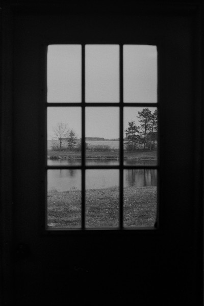 Black and white photo taken from indoors looking at a door what is mostly in shadow but the frame and doorknob can be made out. The door has a 9 pane window and the scene outside is of that of a small pond with some trees on the far end with fields extending beyond under an empty sky. Most of the pond is in the bottom third, the trees in the middle third, and the top third is completely void of features.