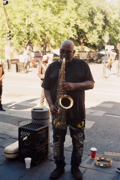 A street musician is standing in on a sidewalk and front of a cross walk with some people and trees which are in the bright sunshine. The musician standing in the shade is an older black man with short gray hair is playing a large shiny saxophone wearing a wearing t-shirt and black jeans with serval colorful patches.