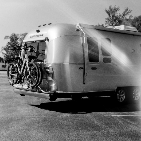 Black and white photo of a classic silverr Airstream camper is show from the back right and only about 70% of the length of the campter is in frame. The camper is in a parking lot under a bright sun and the sunlight is gleaming off the smooth curves of the sides and top. The back of the camper hsa two step-through electric bicycles. The whole vibe is ready for summer adventures. There's a light leak in the photo that looks like a stylistic flare across the right side of the camper. Shot with a Kiev 88 and ArsatC80f28 lens, on Kentmere100, and developed with FX-39II by Shom Bandopadhaya. Licensed under Creative Commons Attribution-NonCommercial-ShareAlike (CC BY-NC-SA).