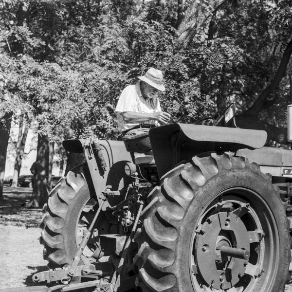 Black and white photo of an old white man wearing a short sleeve button shirt and suspenders with a straw hat is sitting sideways on a big tractor looking rather old-timey. His face is in the shade while everything is baking in the sun, he's looking down at his hands (he's holding a phone that I worked hard to obscure from angling) the rear view mirror shows another angle of his jaw and torso. Only the back half of the tractor is visible with its oversized tires and bit of the engine compartment with a lot of trees in the background. Shot with a Kiev 88 and ArsatC80f28 lens, on Kentmere100, and developed with FX-39II by Shom Bandopadhaya. Licensed under Creative Commons Attribution-NonCommercial-ShareAlike (CC BY-NC-SA).