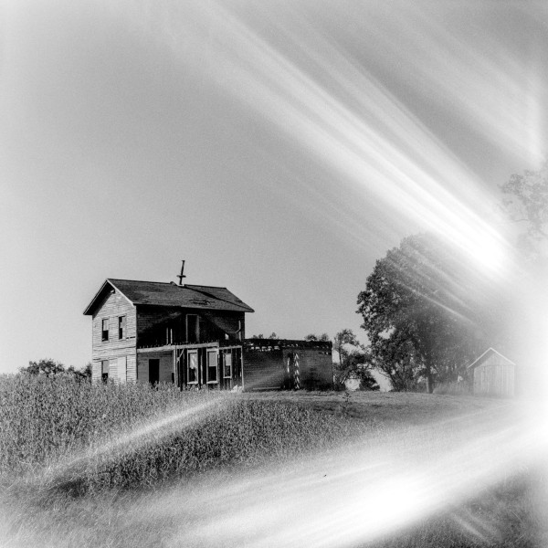 A black and white photo of a dilapidated farm house standing in a field of long grasses. The farm house is two stories with an extension on the side that is a single story which somewhere between unfinished and disrepair. There's a large tree in the background alongside a shed. The entire photo is very heavily affected by light leaks emanating from the middle right and streaking across the frame both up and down in a V shape almost as if destroyed the house and now is sweeping across the landscape. Shot with a Kiev 88 and ArsatC80f28 lens, on Kentmere100, and developed with FX-39II by Shom Bandopadhaya. Licensed under Creative Commons Attribution-NonCommercial-ShareAlike (CC BY-NC-SA).