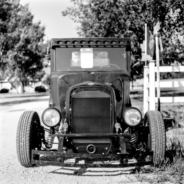 Black and white photo of an old Chevrolet hot rod car parked at the end of a drive way with a partial fence visible to the right. The car is very bare ones with tires mounted on exposed sprints sticking out and big bug eyed head lights and a very flat windshield. At once minimal but classy, a prized possesion of someone who invested a lot of time in it. Shot with a Kiev 88 and ArsatC80f28 lens, on Kentmere100, and developed with FX-39II by Shom Bandopadhaya. Licensed under Creative Commons Attribution-NonCommercial-ShareAlike (CC BY-NC-SA).