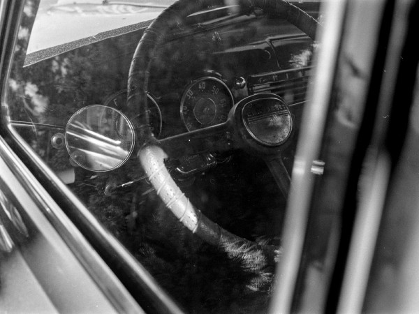Black and white photo of peering into the window of an old car, with the perspective of smooshing your cheek against the window to see inside on a sunny day when the inside is dark. The driver side  window glass is reflecting the round side view mirror and beyond which there is leather bound steering wheel sitting in front of an instrument cluster with round dials showing the speedometer and partial gas tank and other obscured items.