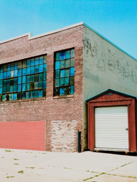 A red shed with a white shutter door and thatched roof is to the right of a larger brick facade building. The side of the brick building is painted yellow but weathered, the front has has a set of large windows with many smaller glass panes. The glass panes are different hues of blue almost like a paint swatch ranging from cerulean to dark gray. The bottom of the building has some boarded up with bricks and concrete blocks painted a deep pastel salmon. Shot with a Mamiya SixVa and Zuiko75f3.5 lens, on HarmonPhoenix200I, and developed with BelliniC41 by Shom Bandopadhaya. Licensed under Creative Commons Attribution-NonCommercial-ShareAlike (CC BY-NC-SA).