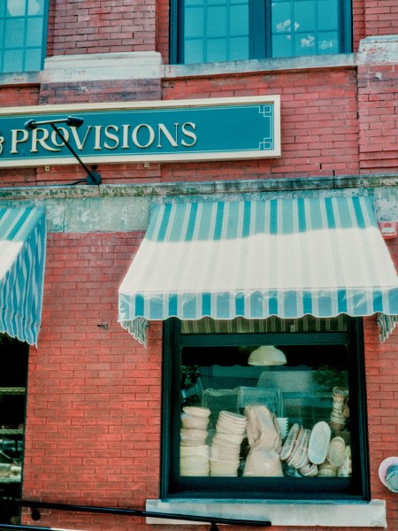 A storefront window with a lot of to-go containers piled up in the window which has a blue and white striped awning partially lit by the sun. The building has a red brick facade with a partially visible sign that is golden lettering on blue background that says "Provisions". The contrast of the different blues, gold, and red are at once vivid but not that colorful. Shot with a Mamiya SixVa and Zuiko75f3.5 lens, on HarmonPhoenix200I, and developed with BelliniC41 by Shom Bandopadhaya. Licensed under Creative Commons Attribution-NonCommercial-ShareAlike (CC BY-NC-SA).