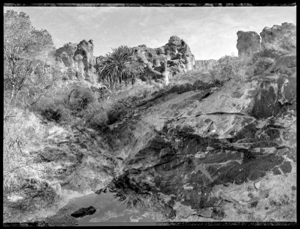 Black and white photo of desert ravine scene with a black border that has a fuzzy internal border as if the image shifted on itself. There are prominent rock faces and a few trees in the top third and a body of water reflecting of the trees and a sky of scattered clouds in the foreground. The rest of the photo the ravine looks natural until you take a closer look and everything is just a bit undefined; fuzzy but not blurry, there but also floating.