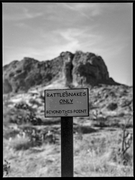 A black and white double expsoure of a desert scene, the film rebate of a 6x4.5 frame can be seen with the black edge getting a small cat eye cut in. The foreground is a prominent sign reading "Rattlesnakes ONLY. Beyond this point." in the middle of the frame mounted on a post. Beyond the sign stretching into the out of focus blurry background is a prominent rock face on the side of a hill on a fairly steep hill. The entire scene is light in bright sunlight with some wispy clouds in the sky. The sign is in the shade but against the bright background making it stick out more.