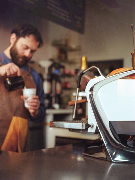 A high end commercial espresso machine is seen in profile in bright white and shiny chrome curves taking up a third of the frame right. As focus falls away to the mid-ground a barista wearing a grown apron, blue shirt, and a thick beard can be seen pouring milk from a pitcher into a cup. The background almost totally blurry is coffee shop standards like tall grinders, shelves with brewing tools. The light is that of a low winter sun, taken a couple months ago, finally found the person again to get permission to post.