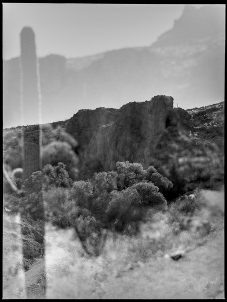 A black and white double expsoure of a desert scene, the film rebate of a 6x4.5 frame can be seen with the black edge getting a small cat eye cut in. The more prominent exposure is a distant rock out on a hillside against a stark sky which is starting to show the other exposure, another shifted over and titled desert horizon. Below the rock out crop are massive trees with their canopy brightly lit and the trunks plunging into darkness. The second exposure also starts featuring in the foreground with a tall pillar of a cactus on the left of the frame simultaneously obscuring and highlighting a path into the valley from the other exposure. The hilly horizon and the angle of the cactus make you want to tilt your head slightly to the left to look at the photo..
