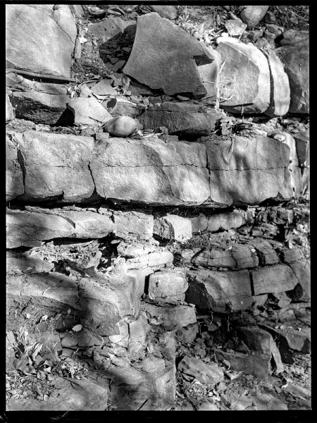 Black and white photo of a rock face lit by the sun casting strong shadows in the nooks and crannies and some shapes from trees as the light is filtered onto the surface. The rocks are layered as if it was put together to form a wall but also very jagged and haphazard in the way they jut out.  There's more texture than there are distinct shades.