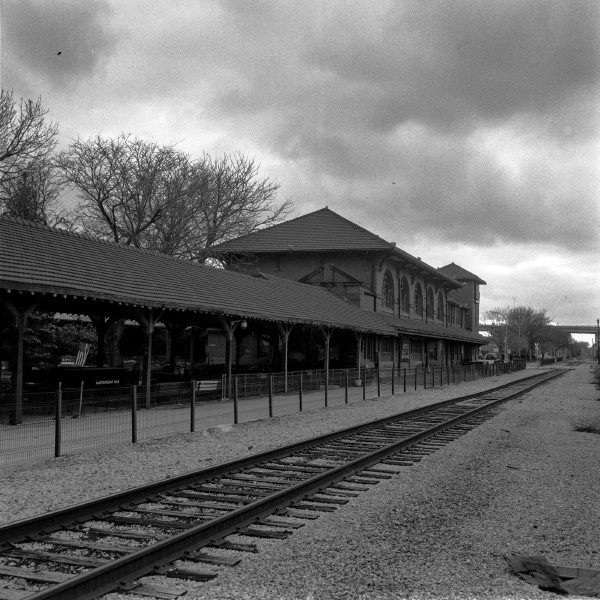 Black and white photo of train tracks extending from the foreground through acrtoss the frame to the middle right on the far side is a building that looks like an old station and there are some gloomy clouds. This place seems like it should be busy but is totally desolate despite being kept up.