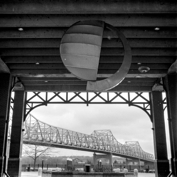Black and white photo of a large archway of an interior space that opens up to the outside. The archway is framing a lattice bridge that stretches from left to right bookend by the brick pillars that are supporting the interior ceiling. The ceiling has horizontal beams from which a sculpture is hanging which is half a sphere on the left and a arcing section of a larger sphere on the right. The entire scene is geometric shapes but each cluster is distinctly different than the rest.