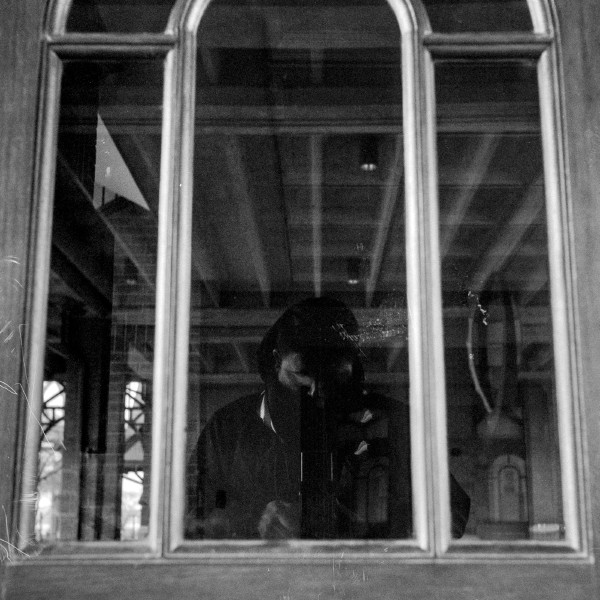 Black and white photo of a large three vertical-pane window which is reflecting the silhouette of a person and the interior of a large open space with exposed ceiling beam. The silhouette is looking down and the vague shape of the top of a Twin Lens Reflex camera can be barely made out if one knows what they're looking at. The bottom of the frame has a lot of scratches adding to the analog blemish look and feel.