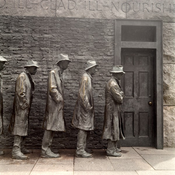 Color photo of life size bronze sculptures in front of a black brick facade with a black door on the right end of the frame. The five figures are facing towards the door, they're all of different heights but dressed similarly in long trench coats and slight variations of a trilby style hats. The expressions of the statues are gaunt, most have their hands in their coat pockets the one in the front of the line has his arms crossed. The bronze statues have the greenish patina except for the shoulders and sleeves where people have touched them, almost as if in support for their plight as if lending strength to them to stay standing in the bread line despite the inclement times and weather. The writing above is mostly cutoff except for the word "NOURISH".