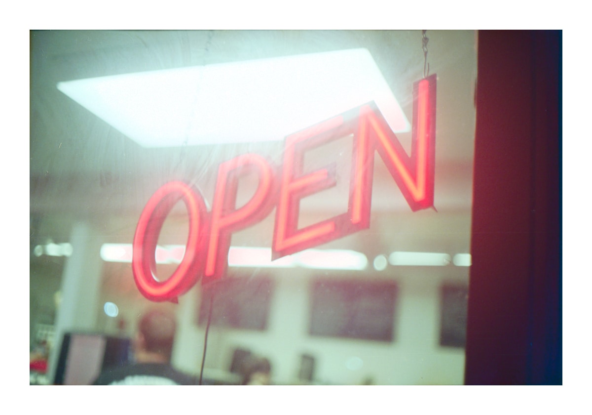 Color photo of a neon sign reading OPEN and an overexposed fluorescent overhead light is visible in the background along with the head and shoulder of a person inside the store window.